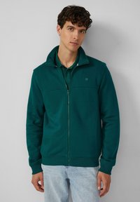 Grüne Kapuzenjacke mit hohem Kragen, aus weichem Stoff gefertigt, mit einem Frontdesign, seitlichen Taschen und gerippten Bündchen.