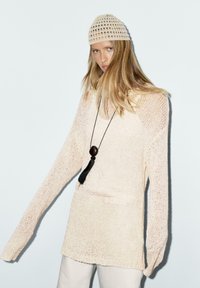 Maglione in maglia beige chiaro con maniche lunghe, scollo ampio e vestibilità morbida. Presenta un motivo testurizzato e una collana con nappina nera.