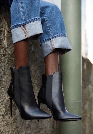 Jambes portant un jean bleu retroussé et des bottines noires à bouts pointus avec talons aiguilles, appuyées contre un mur en pierre à l'extérieur.