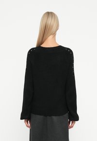 Sweater preto de malha com mangas longas e bainha canelada. Apresenta pequenos tachas ao longo da costura do ombro para um detalhe adicional. Textura suave.