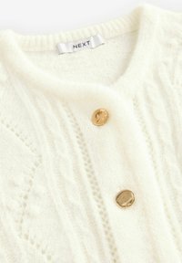 Cardigan de culoare crem cu un design tricotat în cablu, textură moale și două butoane aurii în față. Eticheta citește "NEXT."