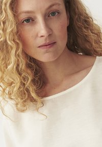 White, linen-top with a rounded neckline; model has wavy, blonde hair and natural skin tone; neutral background enhances shirt's simplicity.