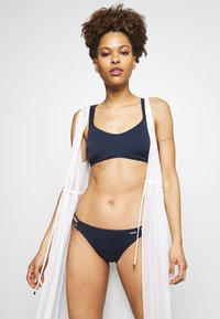 Navy bikini set with a fitted top and low-rise bottoms, featuring three side straps. Lightweight white cover-up with drawstrings.