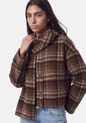 Jeune femme aux longs cheveux foncés portant une veste en tartan marron à boutons et un jean bleu clair, tenant le col de la veste.