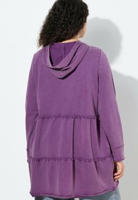 Žena s kudrnatými vlasy, která má na sobě volný fialový top s dlouhými rukávy a kapucí, s nařasenými několika řadami švů, zobrazená zezadu.