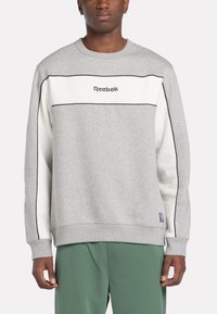 Homme portant un sweat-shirt Reebok gris et blanc avec des bordures noires, associé à un pantalon vert, se tenant devant un fond uni.