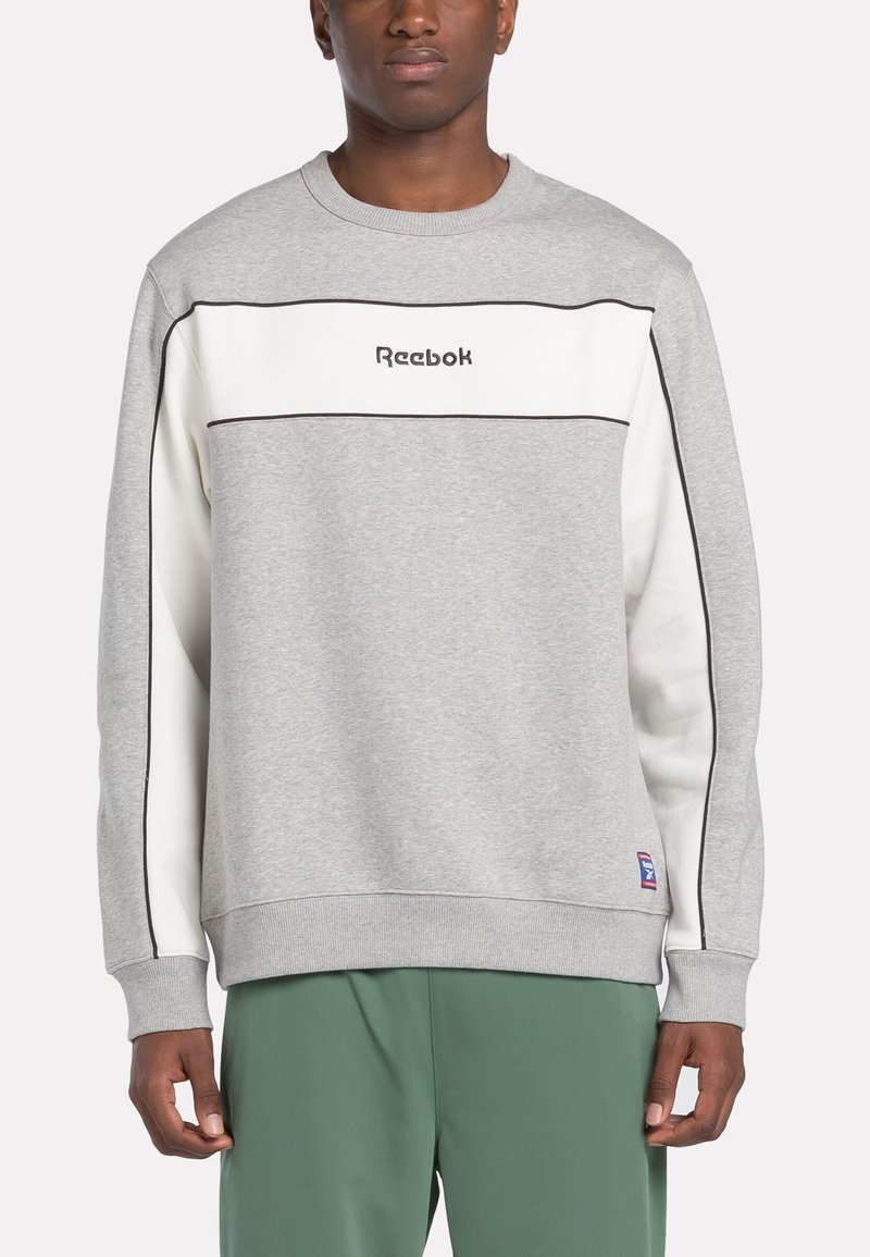 Homme portant un sweat-shirt Reebok gris et blanc avec des bordures noires, associé à un pantalon vert, se tenant devant un fond uni.