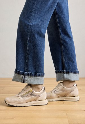 Voeten in beige sneakers met gouden ritsdetails, gecombineerd met lichtbruine sokken en opgerolde blauwe jeans, staand op een houten vloer.