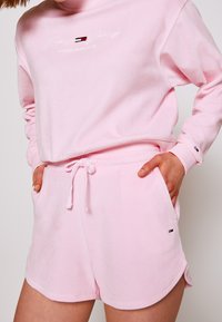 Felpa di cotone rosa con colletto alto e logo ricamato; abbinata a pantaloni corti coordinati con elastico in vita e tasche laterali.