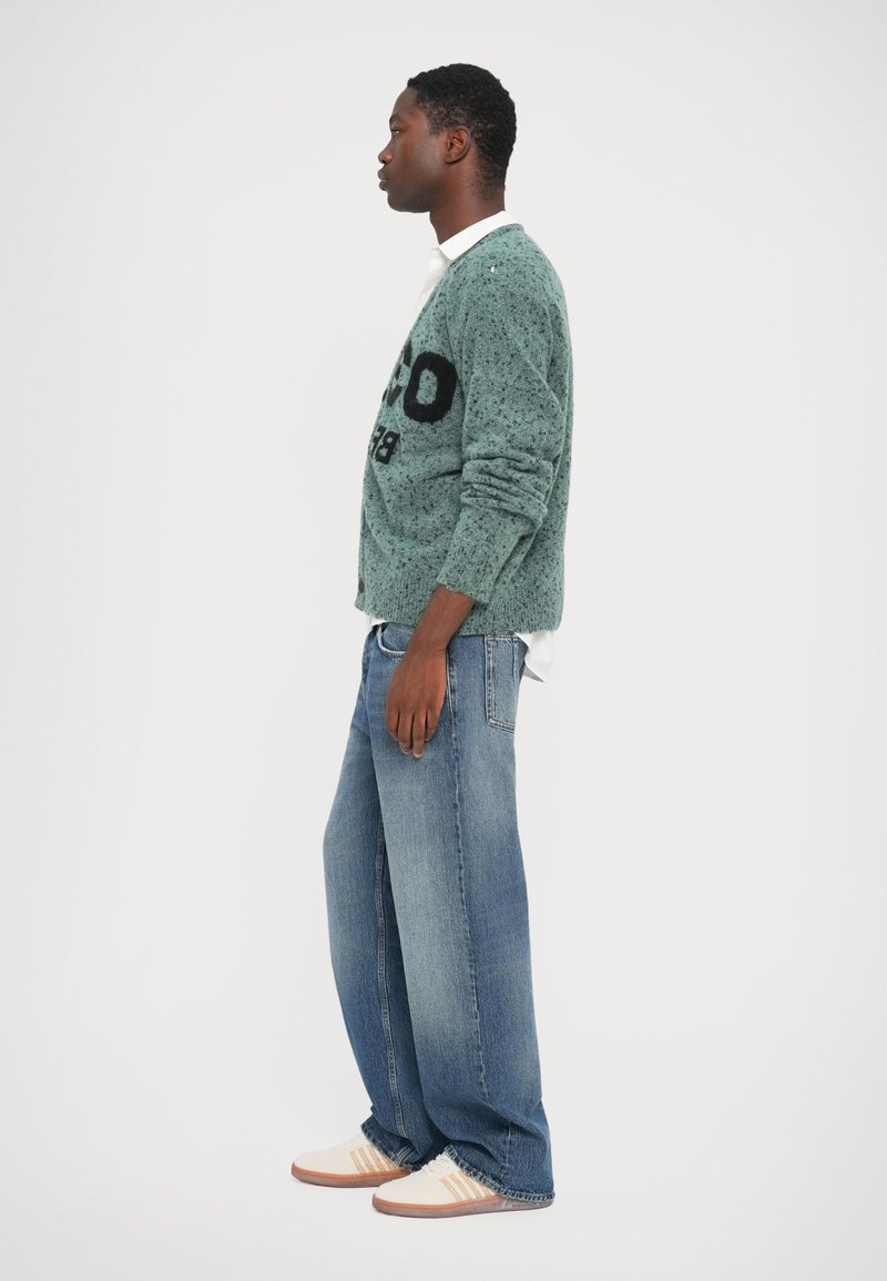 Green speckled sweater with black text, blue wide-leg jeans, and beige shoes with stripes. Model stands in profile against a white background.