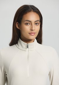Young woman with medium brown hair wearing a cream-colored, long-sleeve top with a front zipper collar, looking softly at the camera.