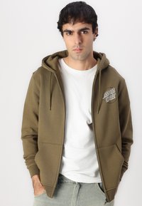Olivnozelena zip-up kapuca z določenimi žepi spredaj in vrvicami, ki ima na levem prsnem delu krožno oznako 'Santa Cruz'. Material mešanice bombaža.