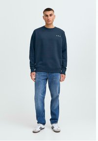 Námornícky crewneck sweatshirt s bielym textom "A.R.C.", mäkká tkanina, uvoľnený strih, kombinovaný so svetlomodrými rovným strihom džínsami a bielymi teniskami.