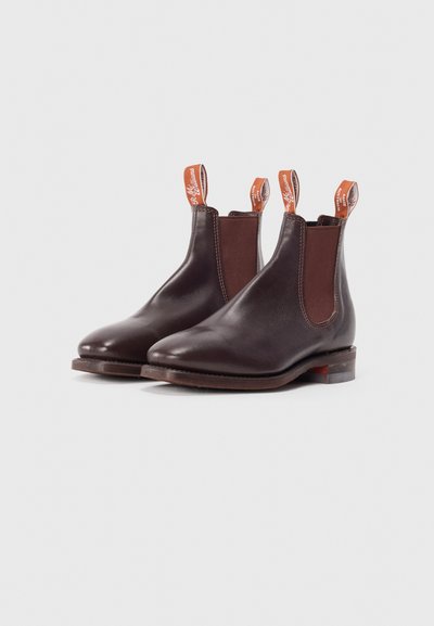 Botine Chelsea din piele maro, cu panouri elastice pe lateral, tălpi din cauciuc și tab-uri de tragere contrastante de culoare portocalie pentru o utilizare ușoară. Textură netedă și vârf rotunjit.