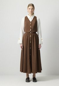 Vestido marrón, de corte A, a la altura de la rodilla, con un cuerpo ajustado y grandes botones, combinado con una camisa blanca de cuello, de manga larga, que exhibe texturas suaves.