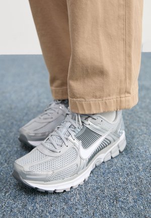 Încălțăminte sport gri cu partea superioară din plasă, accente sintetice netede și logo alb; asortată cu pantaloni khaki, așezată pe o suprafață de covor albastru.