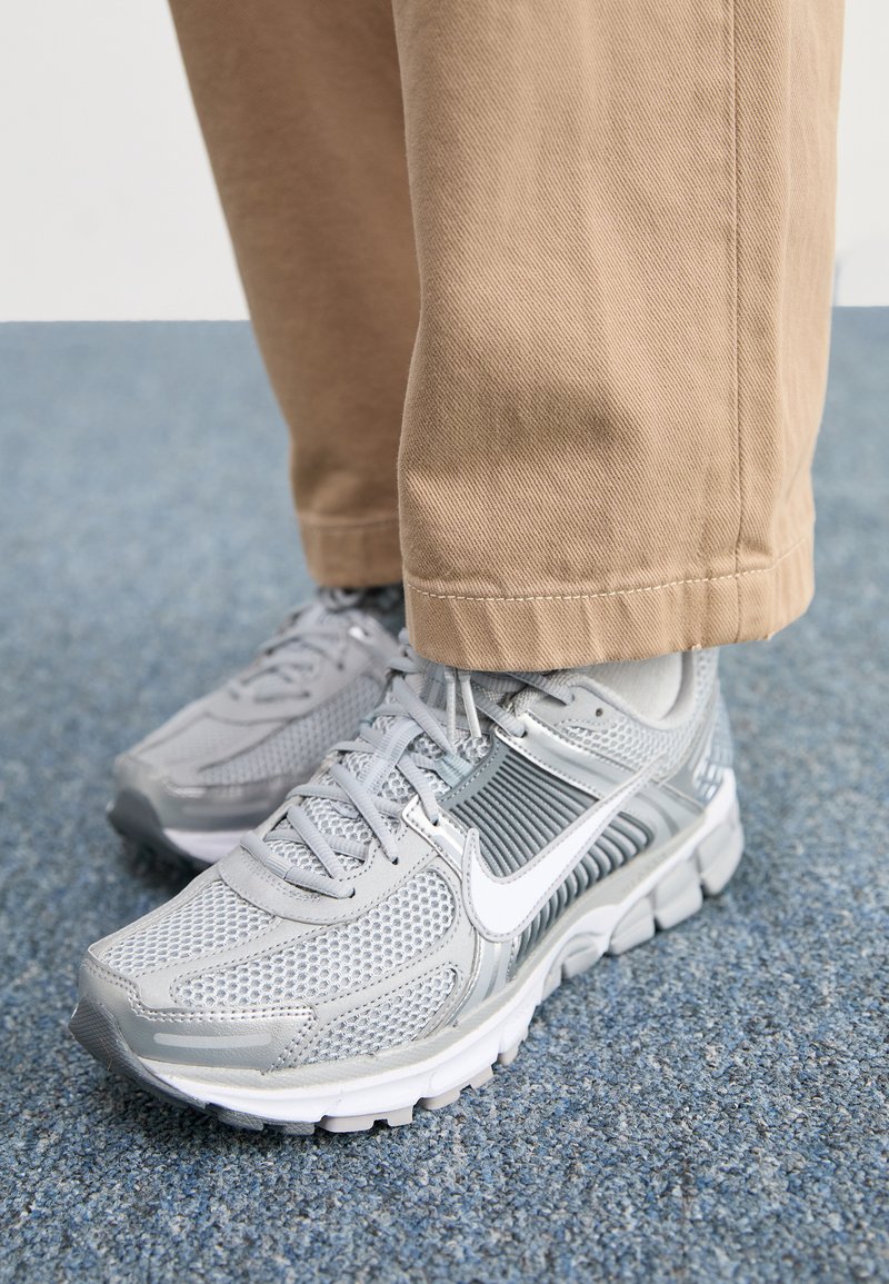 Zapatillas deportivas grises con parte superior de malla, acentos sintéticos lisos y logo blanco; combinadas con pantalones caqui, sobre una superficie de alfombra azul.