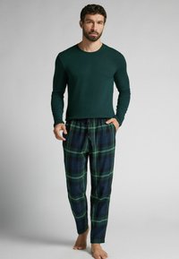 Tmavě zelený top s dlouhými rukávy spárovaný s kalhotami s kostkovaným vzorem v modré a zelené barvě, které mají stahovací pas a přiléhavý střih. Tkanina vypadá měkce.