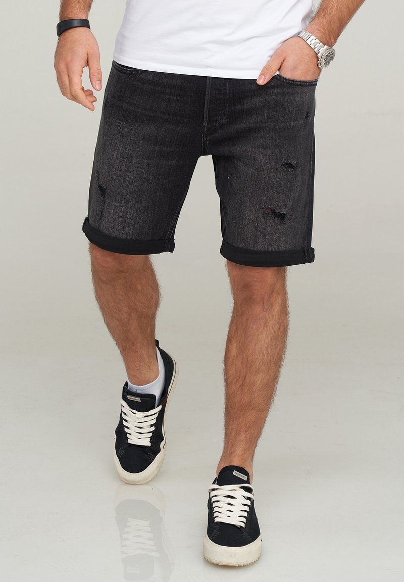 Pantalones cortos de mezclilla negra con dobladillos enrollados, detalles desgastados y un ajuste ceñido. Lleva unos zapatillas negras y calcetines tobilleros blancos.