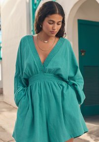 Robe bleu sarcelle avec un décolleté en V profond, taille cintrée et manches amples. Tissu texturé et léger qui crée une coupe décontractée avec des poches latérales.