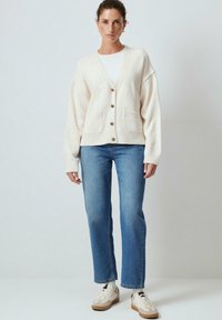Lichtbeige gebreide cardigan met een V-hals, twee voorzakken en grote knopen. Gepaard met blauwe rechte jeans en witte sneakers.