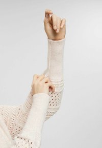 Pull tricoté rose pâle avec poignets côtelés et motifs ajourés sur les manches, mettant en valeur un tissu texturé et des détails complexes.