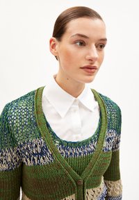 Strickjacke in Grün, Blau und Creme mit offener Webstruktur und Knopfverschluss; über einem weißen Hemd mit Kragen getragen.