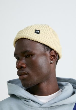 Berretto lavorato a maglia giallo con una texture a coste e un piccolo logo nero, indossato da una persona con un hoodie azzurro chiaro e un colletto stratificato.