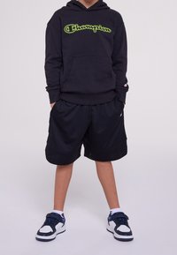 Černá mikina s kapucí se zeleným logem "Champion", doplněná o černé síťované šortky a černé tenisky s bílými a modrými prvky.