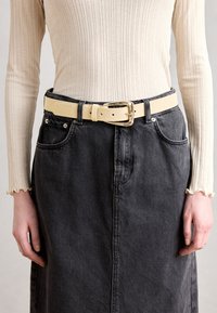 Top de manga larga de canalé color crema, falda de mezclilla negra y un cinturón ancho de cuero beige con una hebilla dorada brillante, llevado a la cintura.