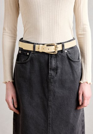 Kremo spalvos kanapinė ilgomis rankovėmis palaidinė, juodi džinsai sijonas ir platus smėlio spalvos odinis diržas su blizgiu auksiniu sagtimi, dėvimas ant liemens.