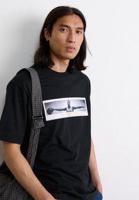 Sort bomulds T-shirt med et grafisk print af en basketballspiller på forsiden, parret med en gråternet skuldertaske.