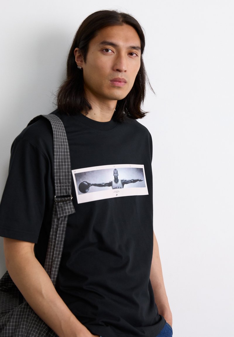 Schwarzes Baumwoll-T-Shirt mit einem grafischen Druck eines Basketballspielers auf der Vorderseite, kombiniert mit einer grau karierten Umhängetasche.