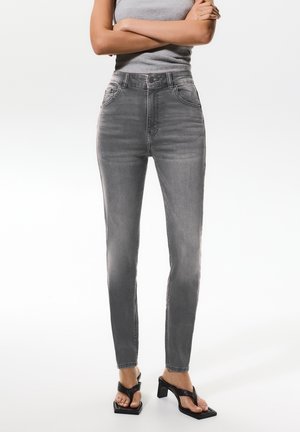 Jeans skinny gri în talie înaltă, cu buzunare frontale și un design decolorat. Asortate cu sandale negre cu toc și un top gri fără mâneci.