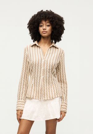 Γυναίκα με σγουρά μαλλιά που φοράει μια μπεζ και λευκή μπλούζα με κουμπιά και σχέδιο ζιγκ-ζαγκ και μια λευκή κοντή φούστα, στέκεται με ουδέτερη έκφραση.