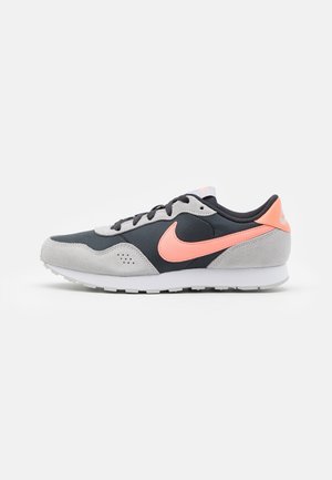 Zapatilla deportiva de perfil bajo en negro y gris con el logo Nike Swoosh en rosa coral y suela blanca, mostrada en perfil derecho sobre fondo blanco.