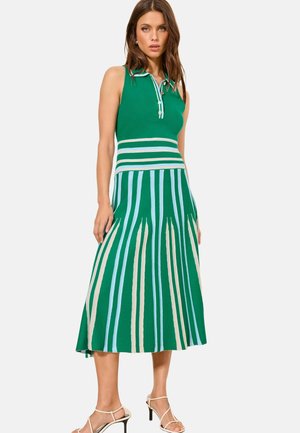 Naine kannab rohelist varrukateta kleiti, mille seelik on valgete, siniste ja beežide vertikaalsete triipudega, seisab ja kannab valgeid kõrge kontsaga sandaleid valgel taustal.