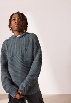 Young person with dreadlocks wearing a textured gray hooded sweater with a zippered chest pocket, standing against a plain light background.