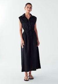 Juodas maxi suknelė su apykakle, išsisegtuku priekyje, dviem krūtinės kišenėmis ir raišteliu juosmenyje. Minkšta medžiaga su lygia tekstūra.