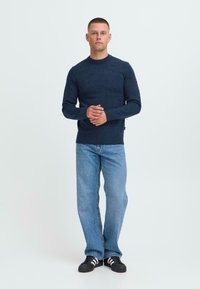 Mann som står mot hvit bakgrunn iført navy genser, lyseblå jeans og svarte sneakers med hvite striper.