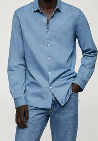 Camicia in denim azzurro chiaro con colletto a punta, maniche lunghe e chiusura frontale a bottoni; presenta una texture liscia e una vestibilità regolare.