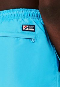 Los pantalones cortos de baño azules presentan una cintura elástica, un bolsillo trasero con cremallera y una etiqueta de tela con acentos en rojo, blanco y azul.