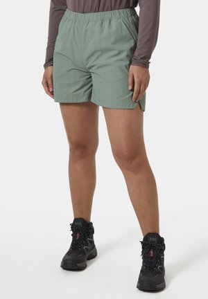 Personne portant un short à taille élastique vert clair, un haut à manches longues gris et des bottes de randonnée noires, debout sur une surface blanche.