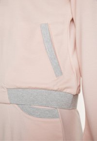 Sweat-shirt et pantalon rose clair avec ceinture côtelée grise et bordures de poche, présentés sur le côté à la taille et aux hanches d'une personne.