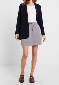 Marineblazer met een enkele pocket, wit geribbeld topje, grijze houndstooth rok met trekkoord in de taille en bruine loafers.
