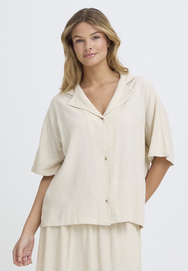 Jeune femme portant une chemise ample beige à manches courtes boutonnée et une jupe assortie, debout devant un fond clair uni.