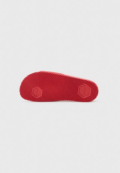 Semelle de chaussure en caoutchouc rouge avec des motifs de bande de roulement hexagonaux et des rainures texturées pour l'adhérence, présentée sur un fond gris uni.