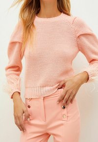 Jasnoróżowy sweter z dzianiny z teksturowanym wzorem i frędzlami przy mankietach, w zestawieniu z dopasowanymi różowymi spodniami o klasycznym kroju z guzikami.