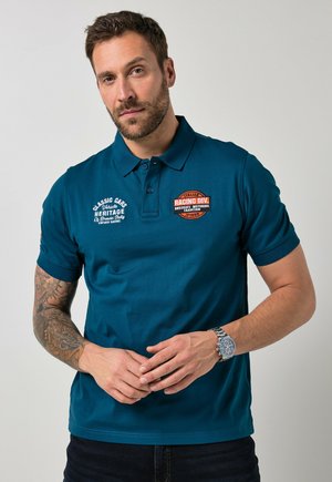 Mężczyzna z brodą i tatuażem nosi turkusową koszulkę polo z naszywkami wyścigowymi, splata dłonie na przodzie, na jednolitym jasnym tle.