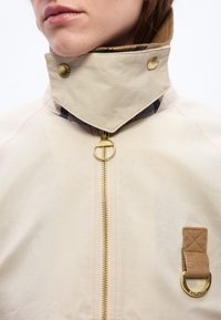 Κοντινό πλάνο για το γιακά μπεζ μπουφάν με φερμουάρ στο χρυσό, χρυσά κουμπιά με πατιλέτα και ταμπά κορδόνι κοτλέ με χρυσό δακτύλιο στο στήθος.